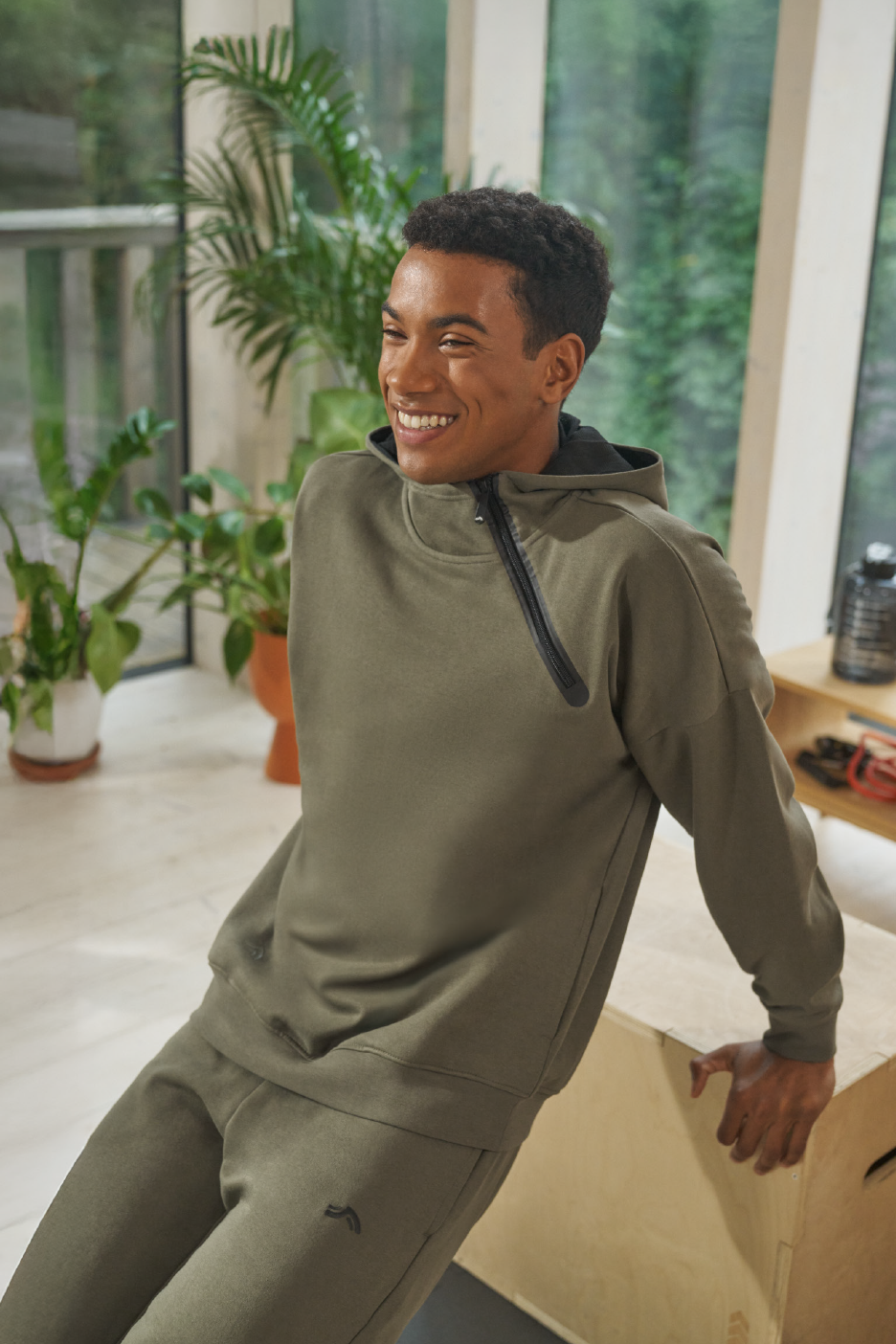 Άνδρας με χακί φούτερ και φόρμα, χαμογελά σε φωτεινό δωμάτιο με φυτά.