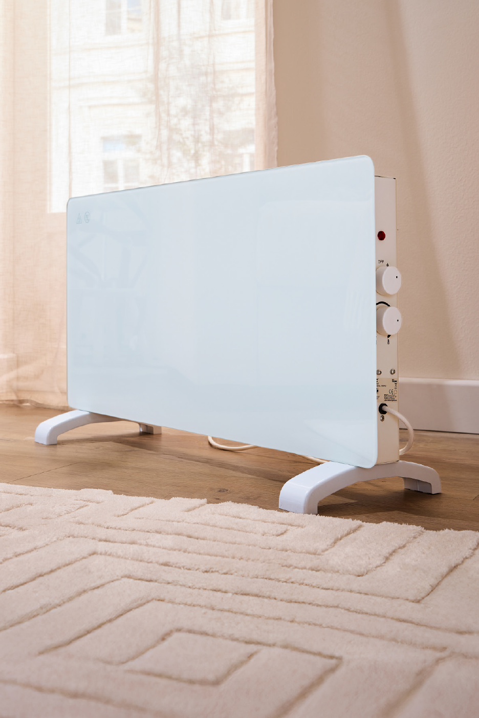 Λευκή γυάλινη θερμάστρα πάνελ με χειριστήρια σε ξύλινο πάτωμα με χαλί, κοντά σε παράθυρο.