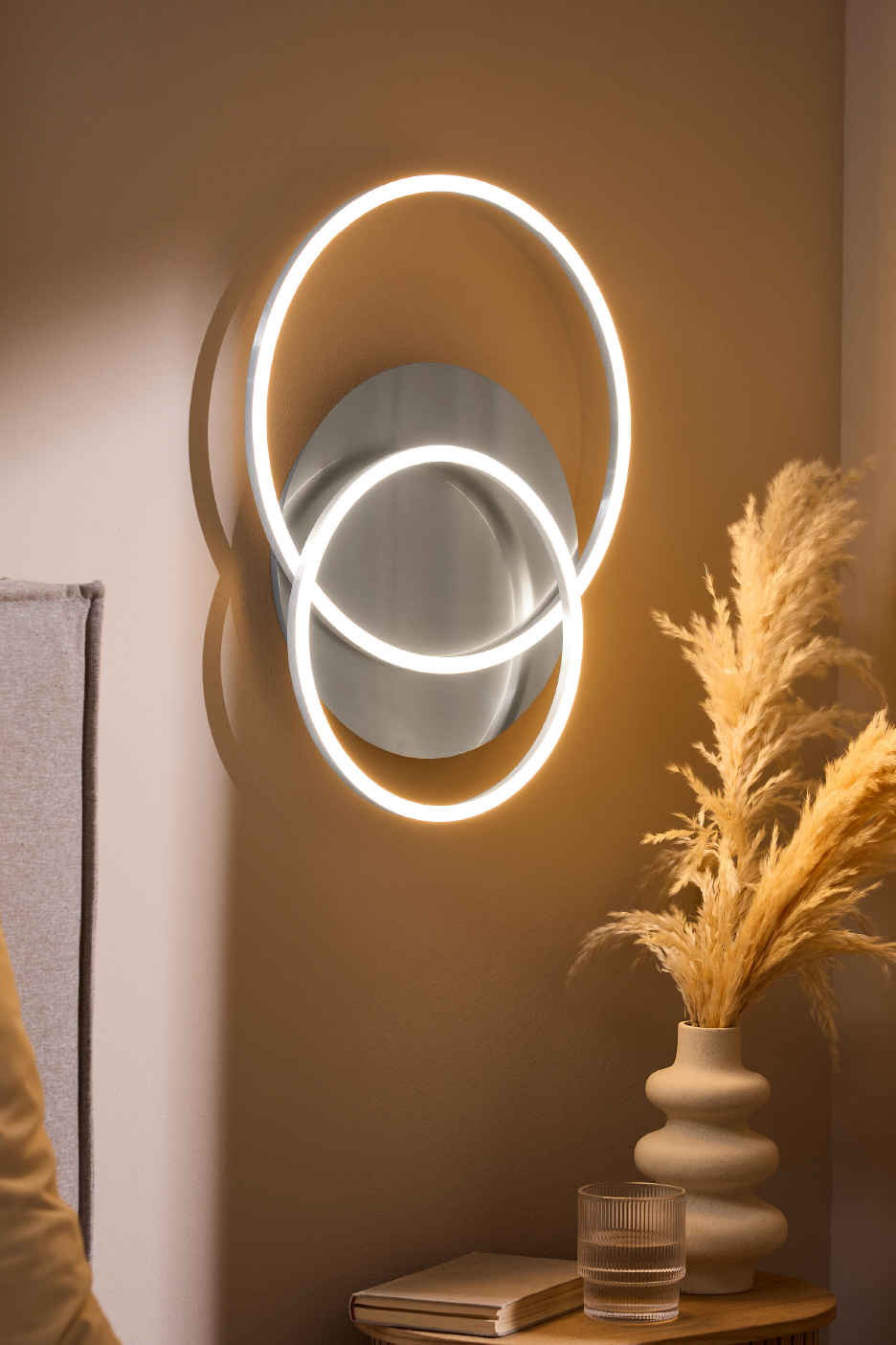 Μοντέρνο φωτιστικό τοίχου LED με δύο συμπλεκόμενους δακτυλίους, που φωτίζει ένα δωμάτιο με ζεστό φως.