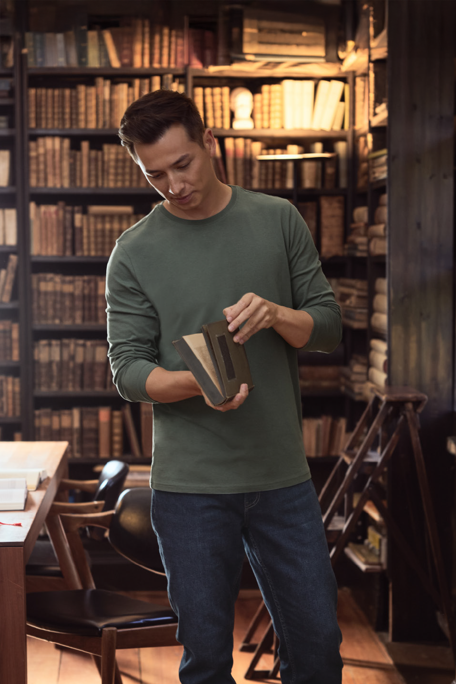 Άνδρας με πράσινη μακρυμάνικη μπλούζα και τζιν κρατάει ένα βιβλίο σε βιβλιοθήκη.