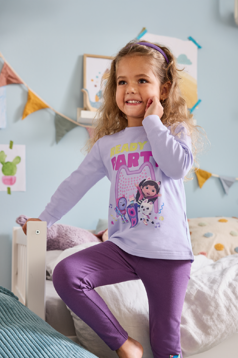 Κορίτσι με μωβ πιτζάμες Gabby's Dollhouse, στέκεται σε κρεβάτι.