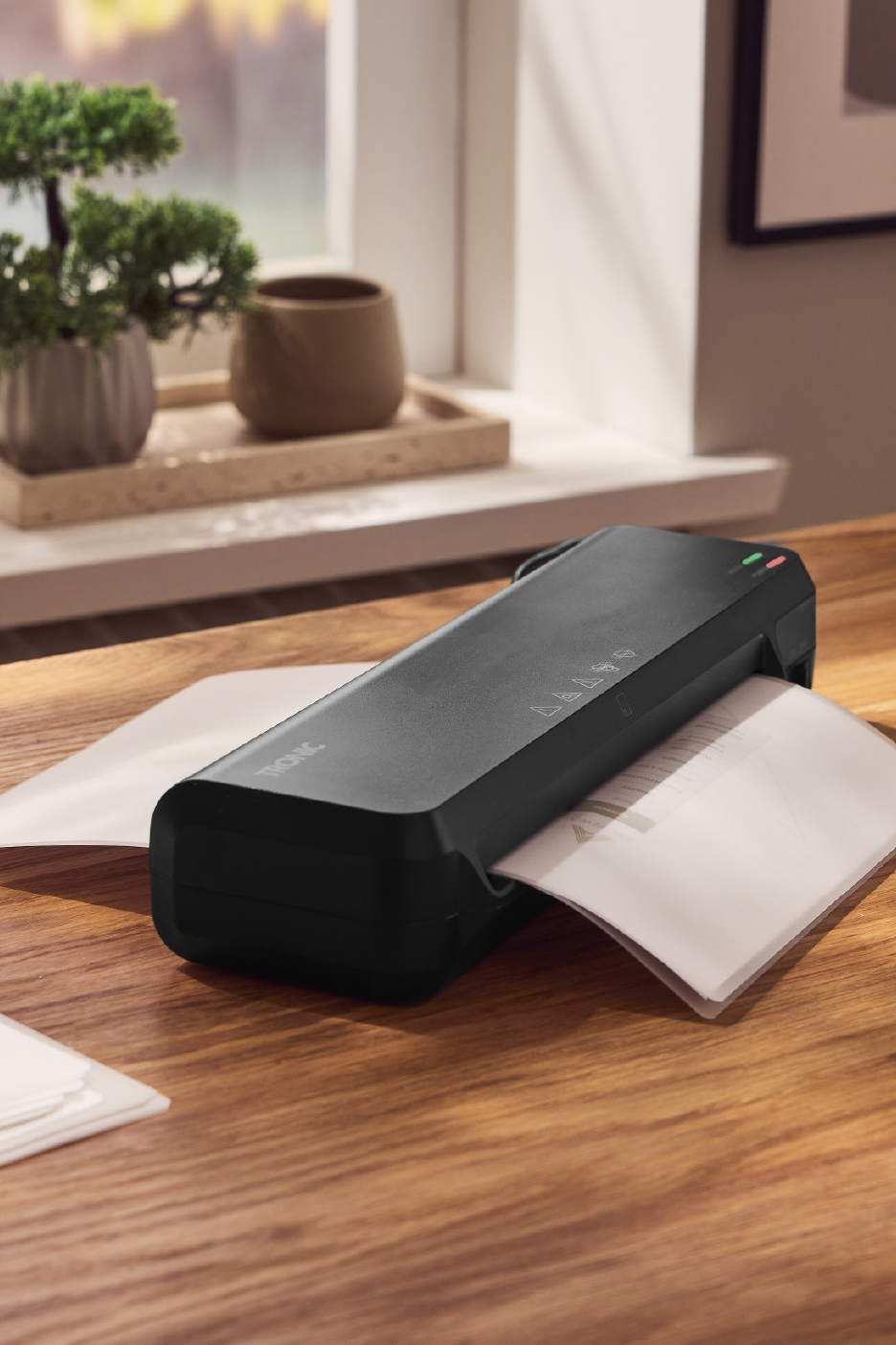 Ένας μαύρος πλαστικοποιητής Tronic σε ξύλινο γραφείο, επεξεργάζεται ένα έγγραφο, με φυτά στο παρασκήνιο.