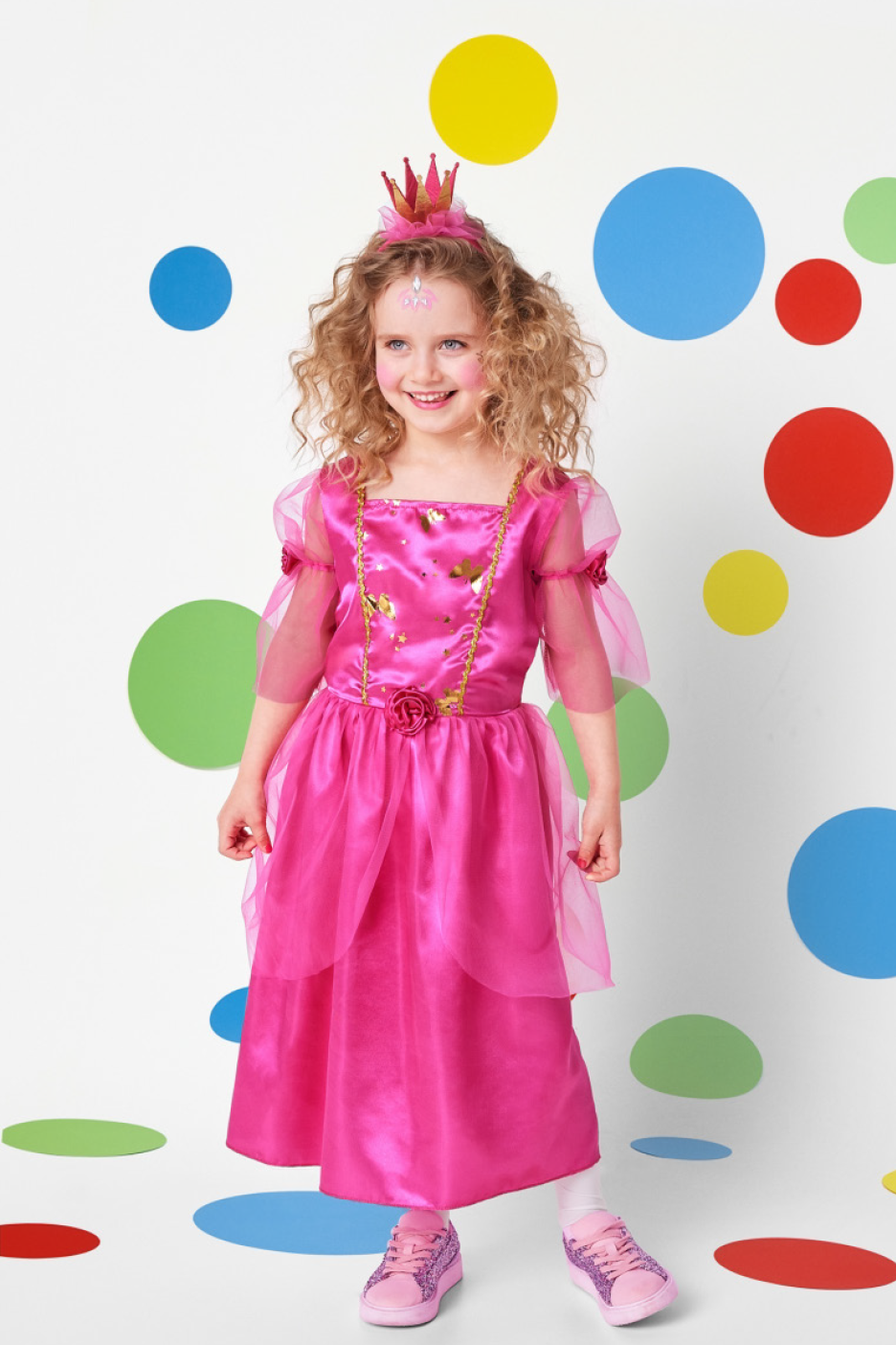 Smiling girl in a pink princess dress and crown, with colorful polka dots background.