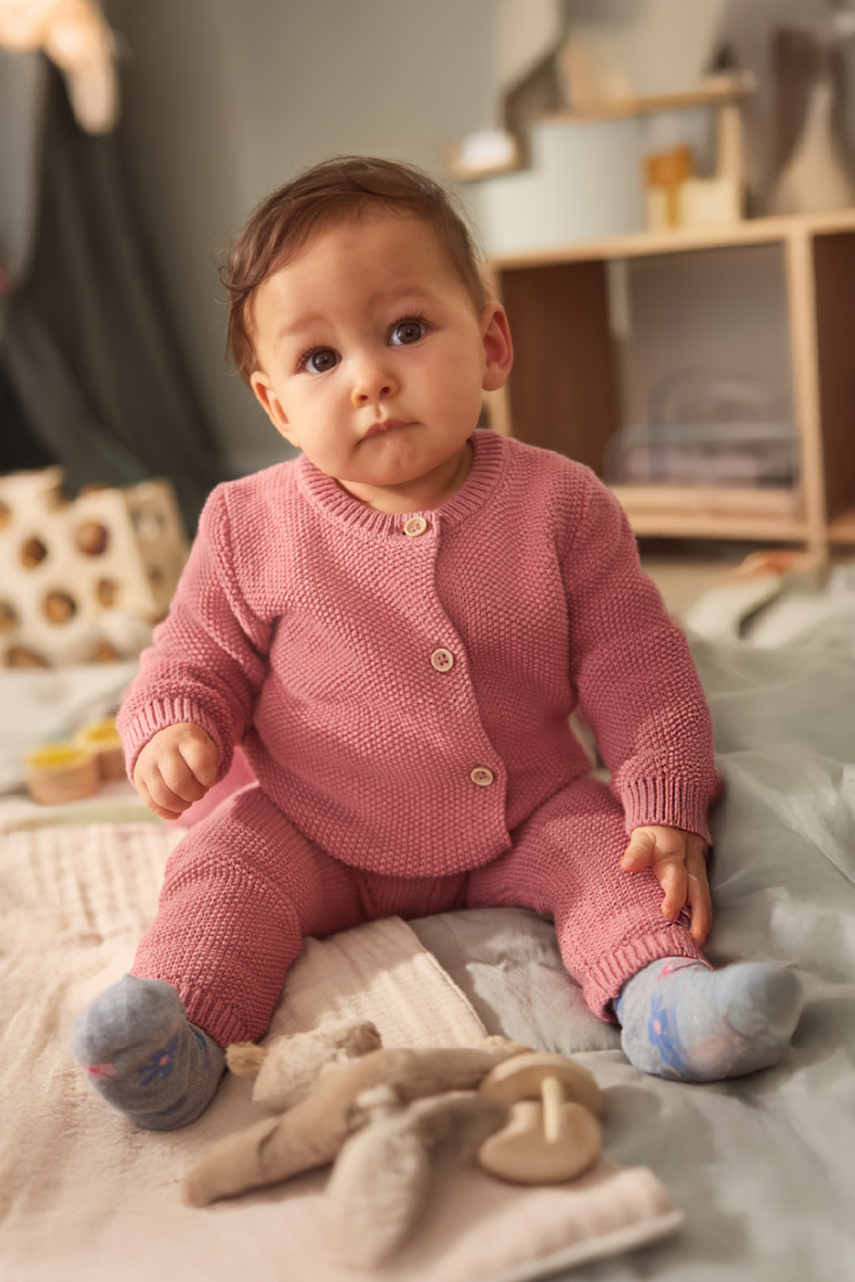 Baby in a pink knitted outfit sitting on a bed with a play tent in the background.