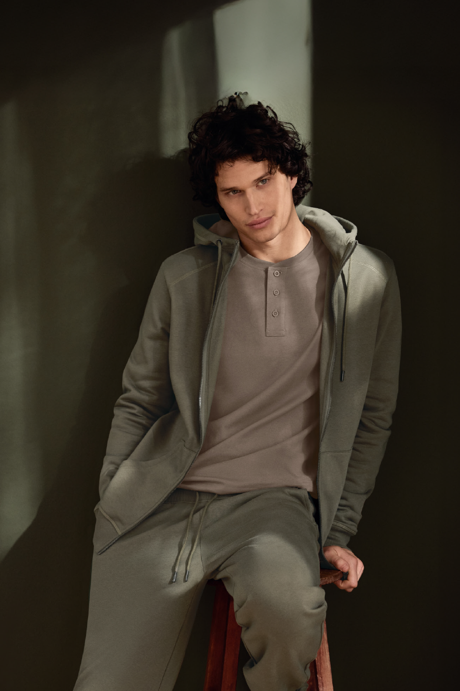 Άνδρας με πράσινο φούτερ, ασορτί φόρμα και μπεζ μπλουζάκι Henley, καθισμένος σε σκαμπό.