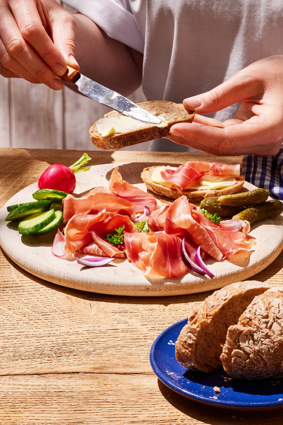 Χέρια αλείφουν βούτυρο σε ψωμί, με πιατέλα προσούτο, τουρσί και ραπανάκια.