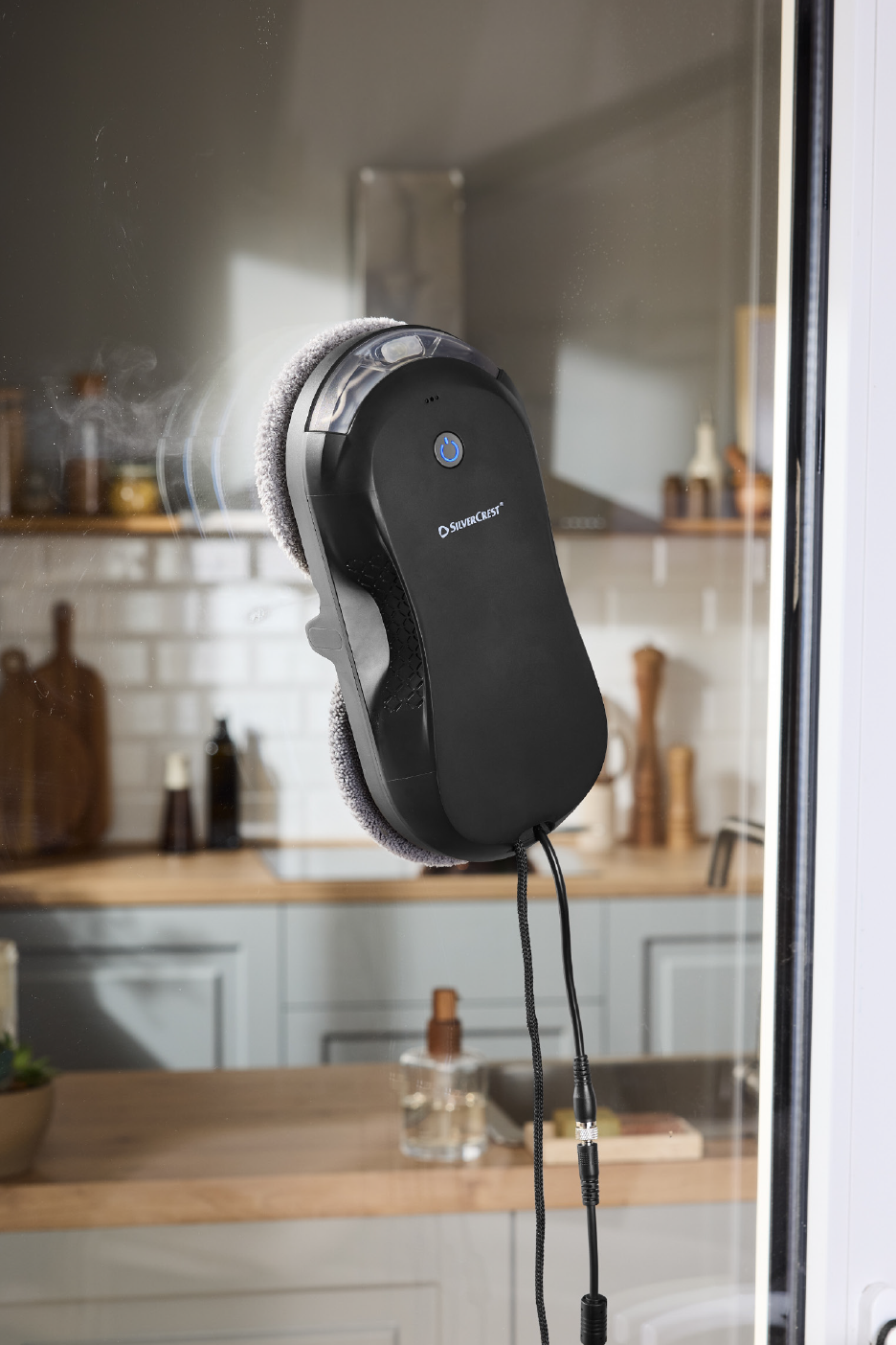 Ρομπότ καθαρισμού παραθύρων σε γυάλινη επιφάνεια, με μια μοντέρνα κουζίνα στο παρασκήνιο.