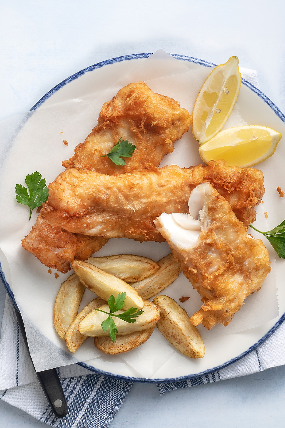 Τραγανό παναρισμένο ψάρι και πατάτες με φέτες λεμονιού και μαϊντανό σε λευκό πιάτο.