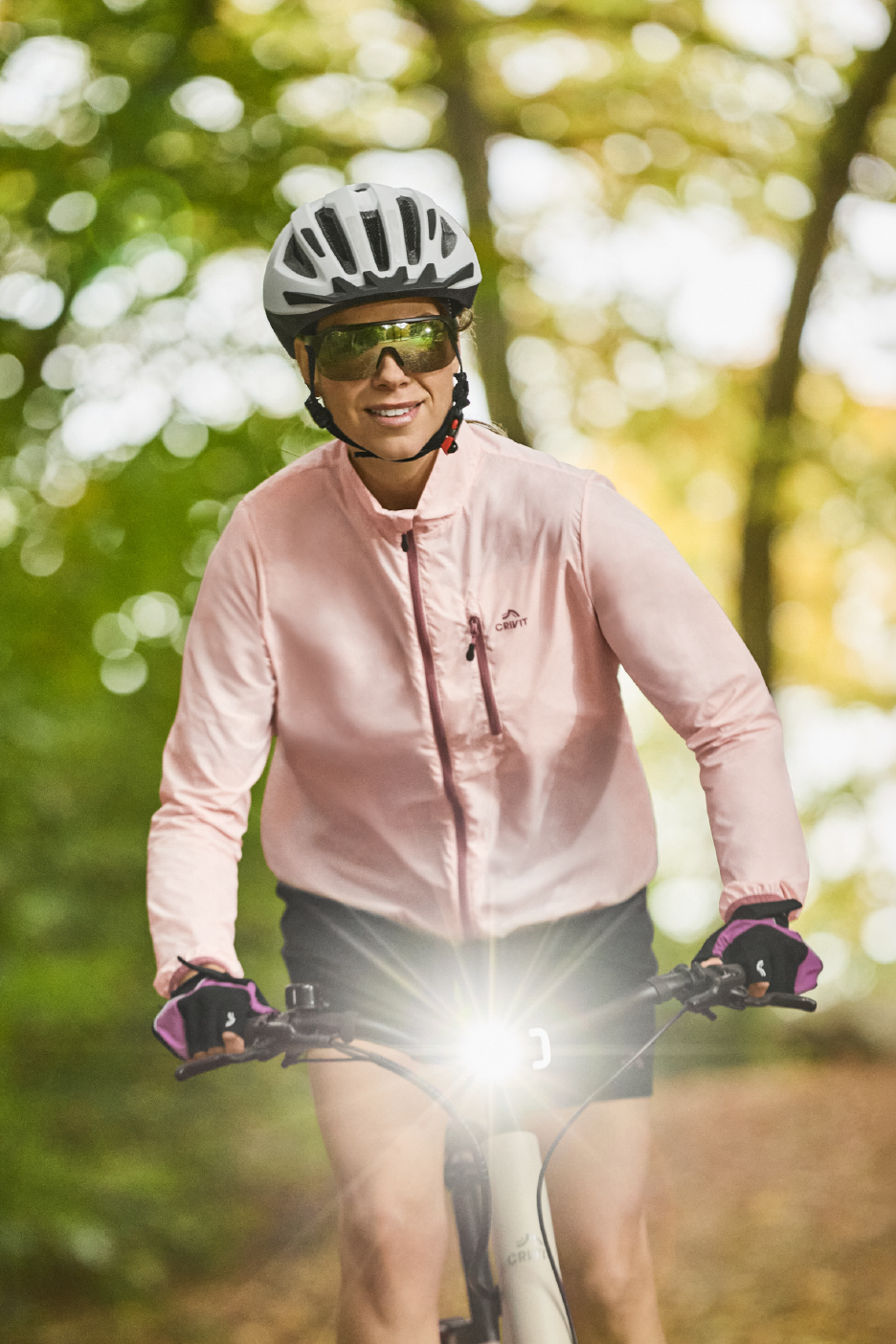 Γυναίκα με κράνος ποδηλάτου, γυαλιά ηλίου και ροζ μπουφάν οδηγεί ποδήλατο με έντονο φως.
