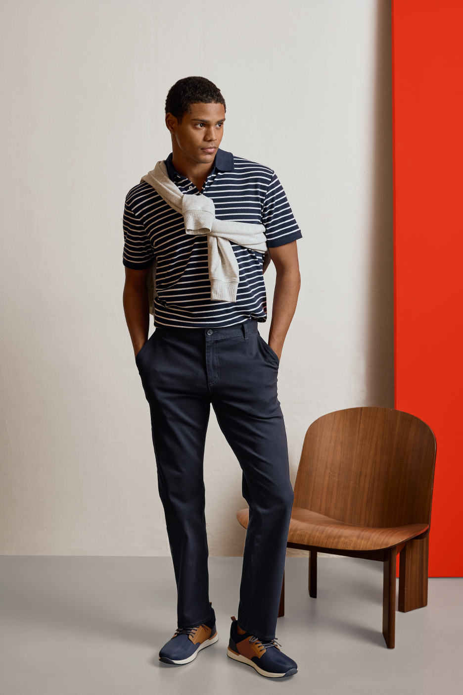 Άνδρας με ριγέ μπλουζάκι πόλο, σκούρο μπλε παντελόνι και αθλητικά παπούτσια, με ένα πουλόβερ στους ώμους.
