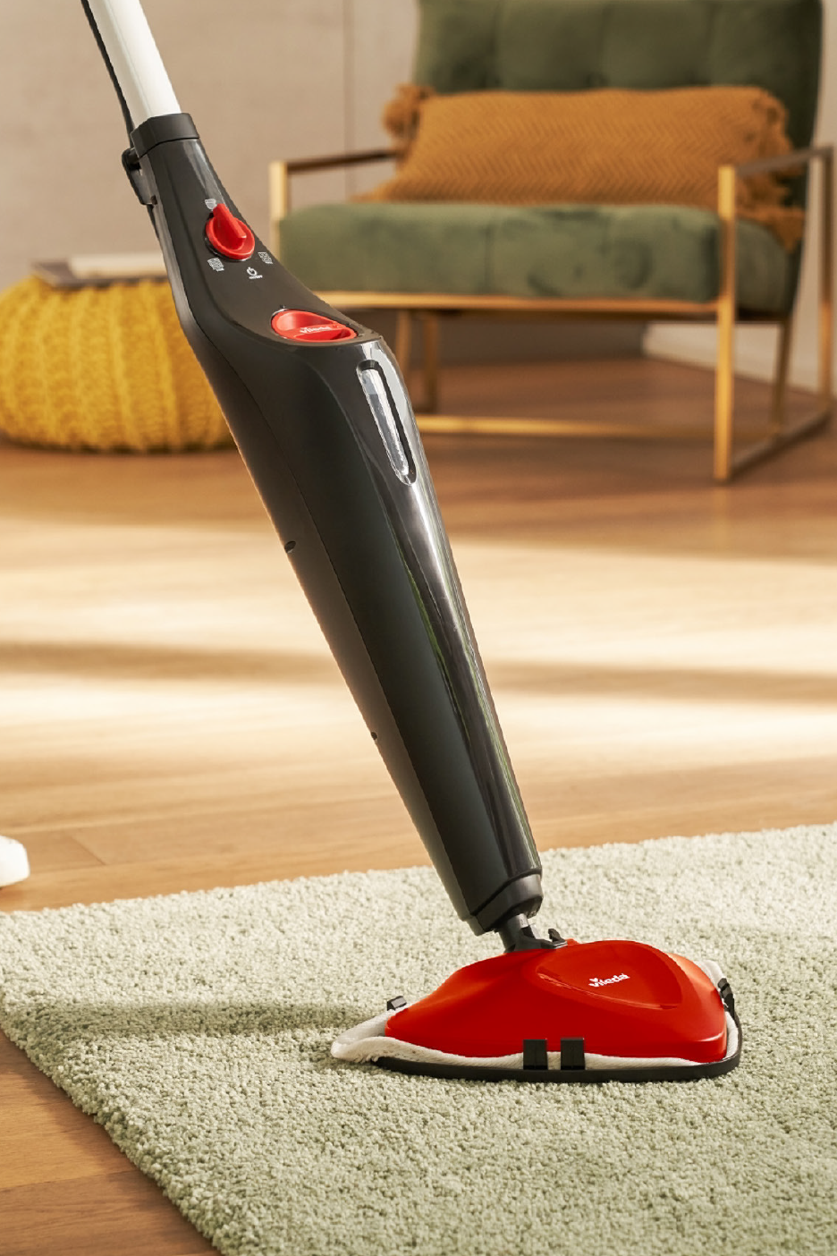 Person cleaning a light green rug with a red and black steam mop on a wooden floor.