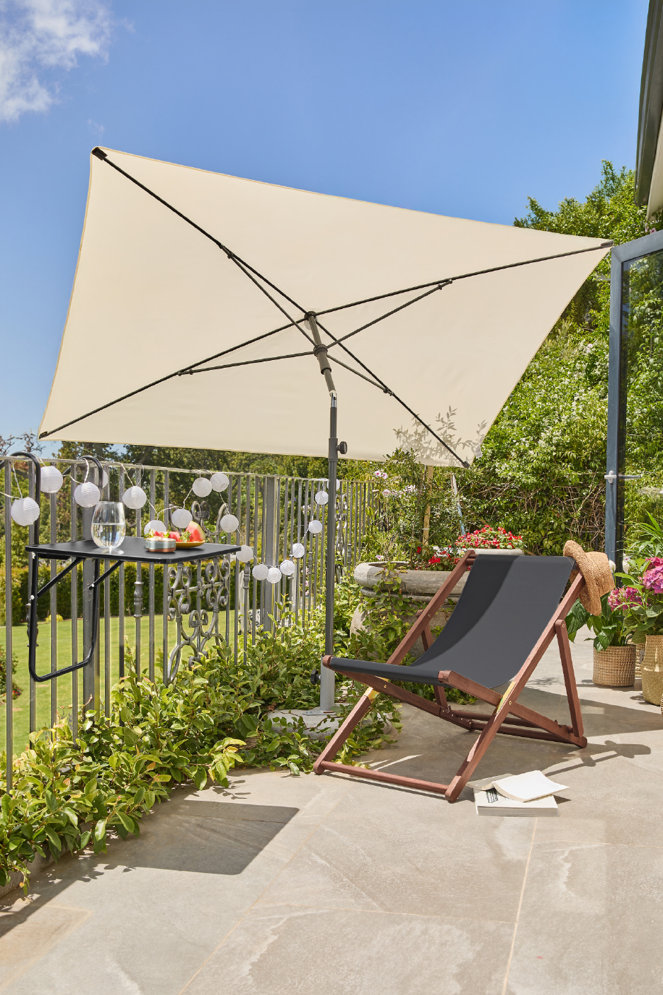 Μπαλκόνι με κρεμ ομπρέλα, μαύρη ξαπλώστρα και ένα μικρό τραπέζι με φρούτα.