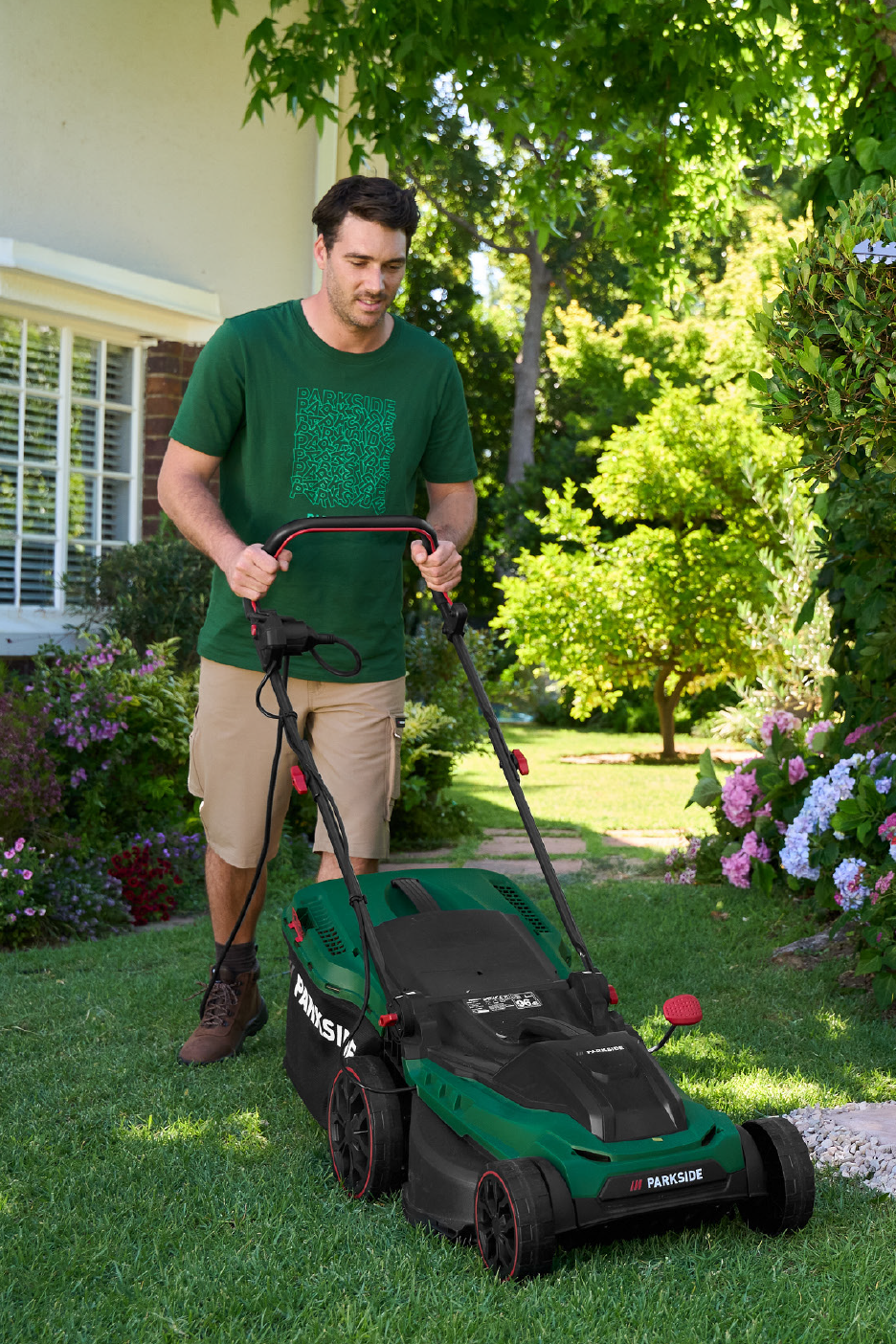Άνδρας με πράσινο μπλουζάκι και σορτς κουρεύει το γκαζόν με πράσινη χλοοκοπτική μηχανή σε κήπο.