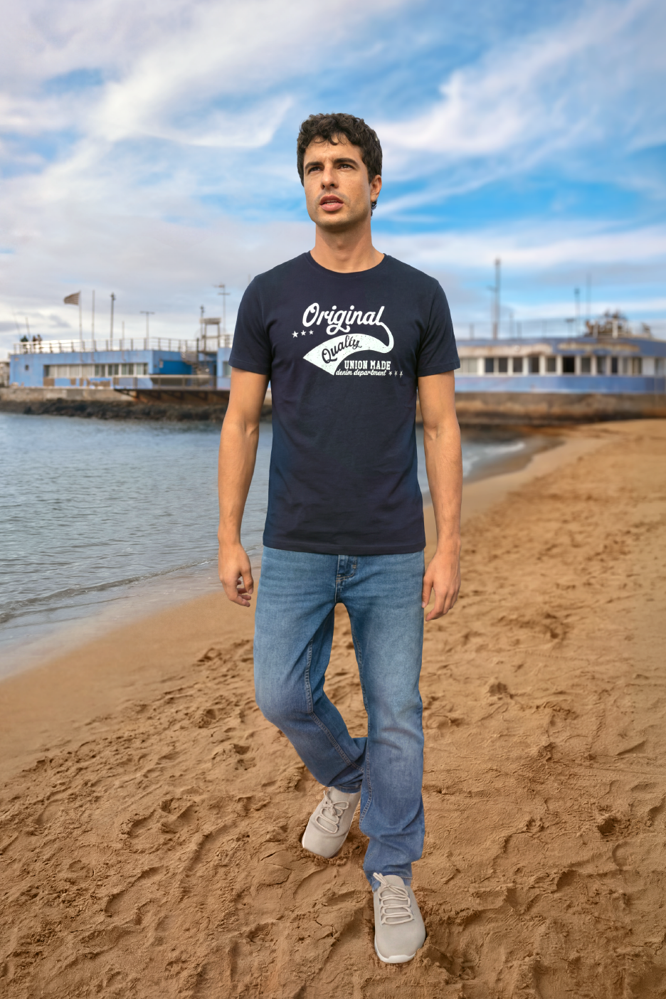 Άνδρας με σκούρο μπλε μπλουζάκι με στάμπα 'Original Quality' και τζιν περπατά σε αμμώδη παραλία.