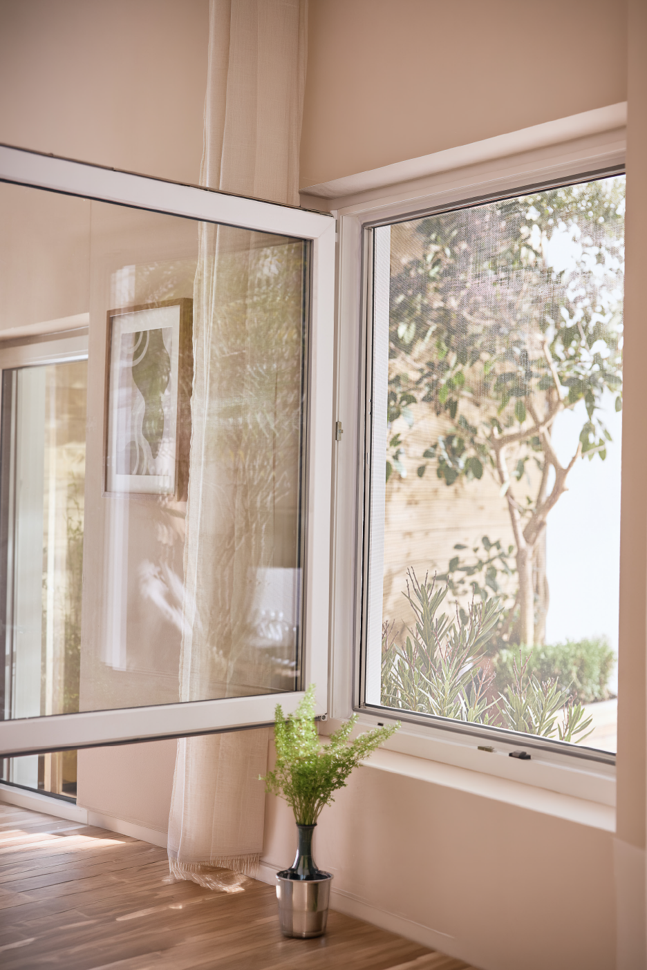 Ανοιχτό λευκό παράθυρο με θέα σε έναν καταπράσινο κήπο, δίπλα σε ένα φυτό σε βάζο.