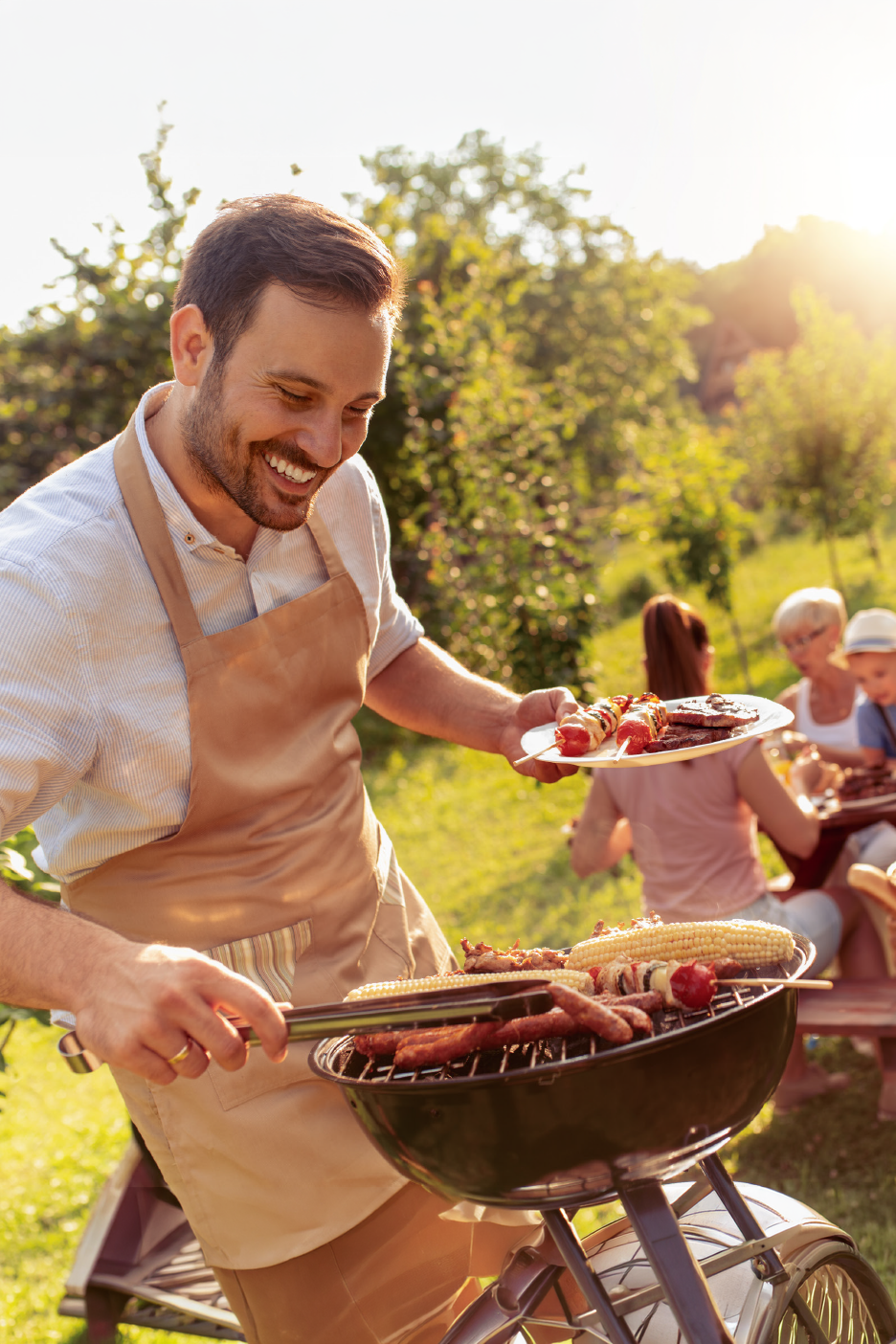 Άνδρας ψήνει λουκάνικα, καλαμπόκι και σουβλάκια, με την οικογένεια να απολαμβάνει μπάρμπεκιου στην αυλή.