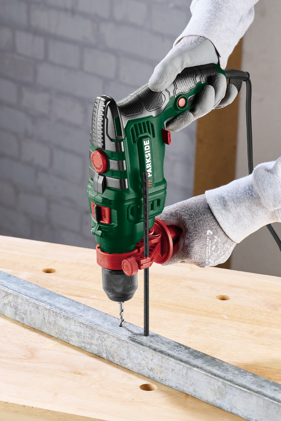 Άτομο τρυπάει μια μεταλλική δοκό με ένα πράσινο και μαύρο ηλεκτρικό δράπανο σε έναν ξύλινο πάγκο εργασίας.