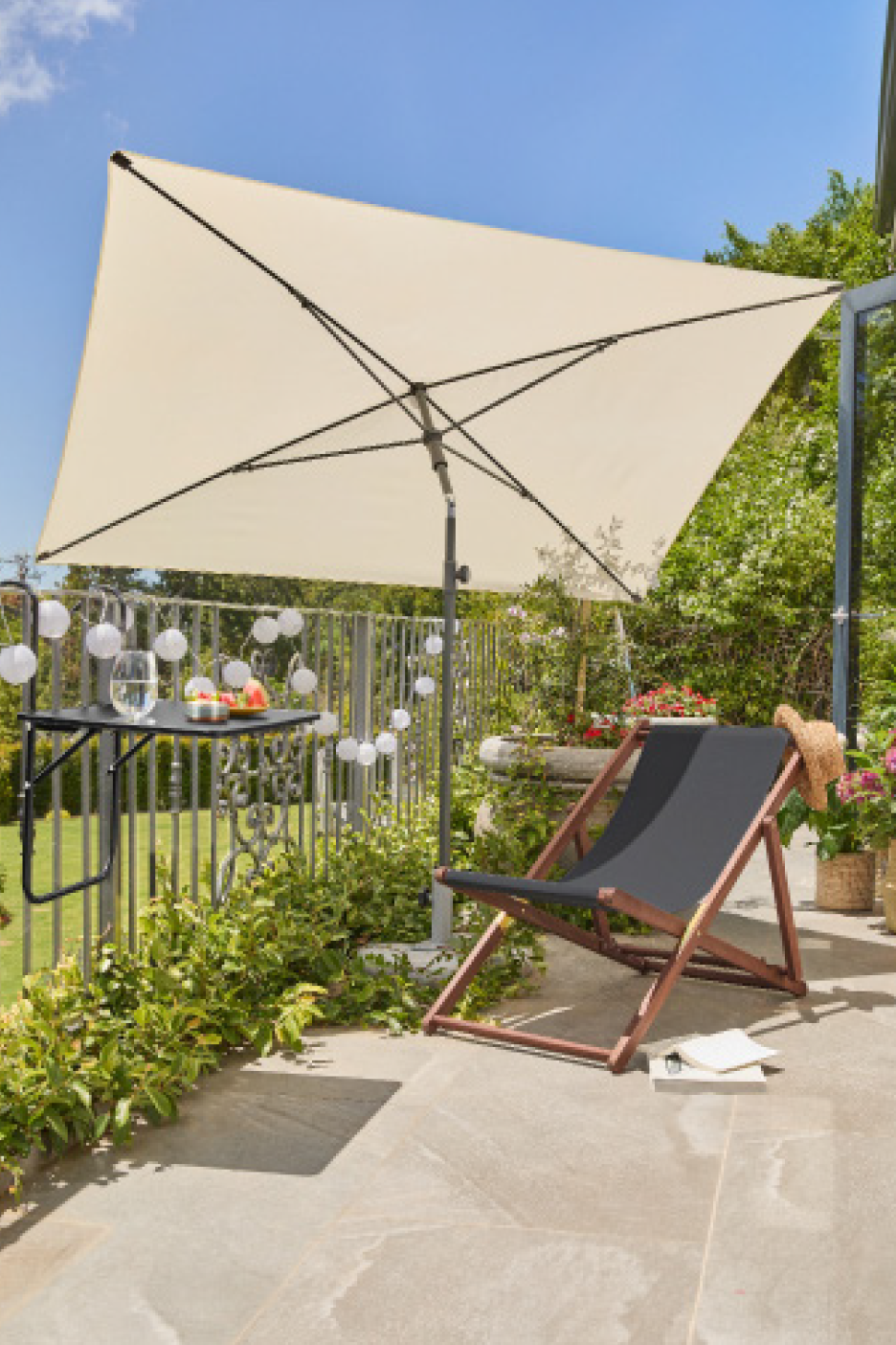 Μπαλκόνι με κρεμ ομπρέλα, μαύρη ξαπλώστρα και ένα μικρό τραπέζι με φρούτα και ποτό.