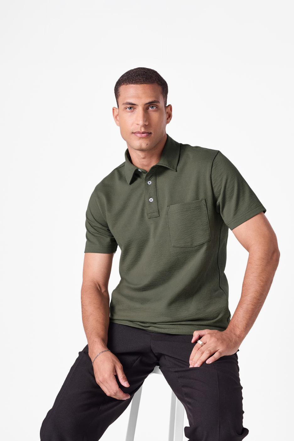 Άνδρας με σκούρο πράσινο μπλουζάκι πόλο και μαύρο παντελόνι κάθεται σε λευκό σκαμπό.