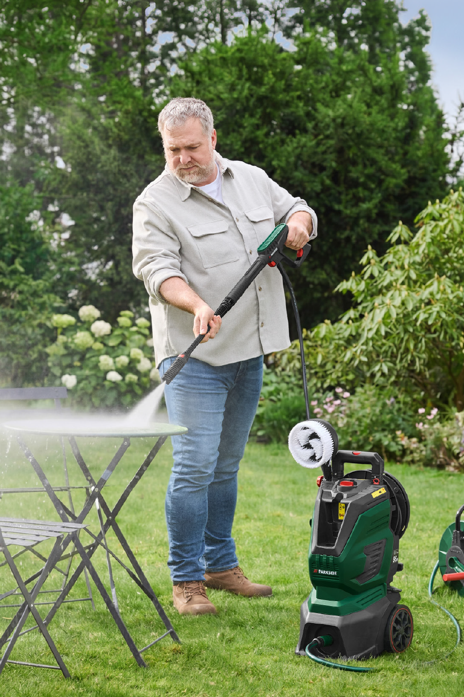 Άνδρας πλένει ένα τραπέζι κήπου με πλυστικό μηχάνημα υψηλής πίεσης σε έναν καταπράσινο κήπο.