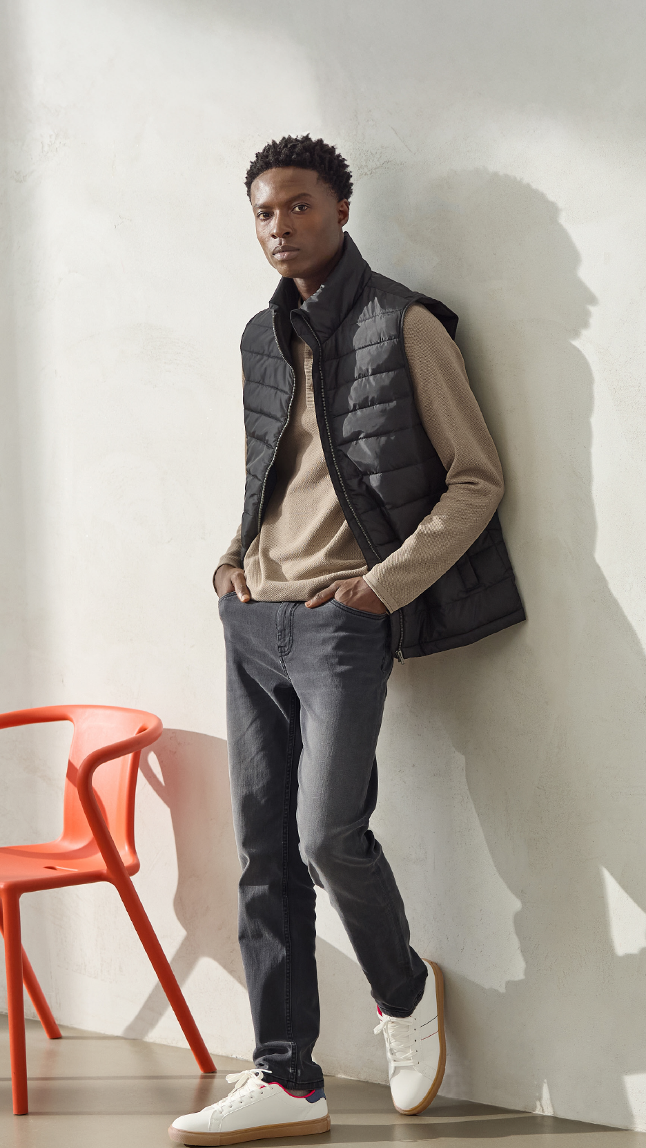Man in black puffer vest and beige long-sleeve shirt, leaning against a wall.