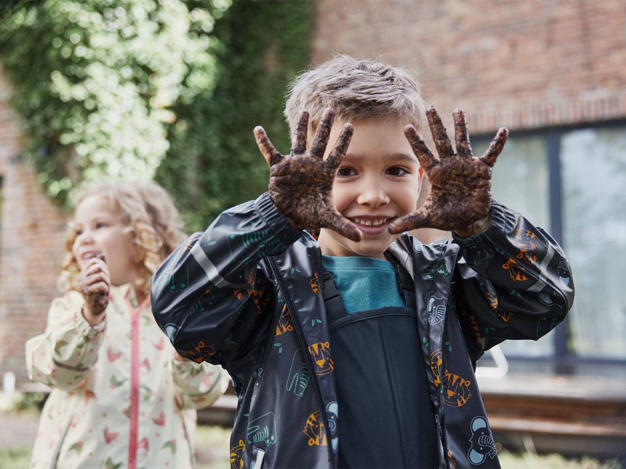 Two children in rain jackets, with muddy hands, smiling outdoors.