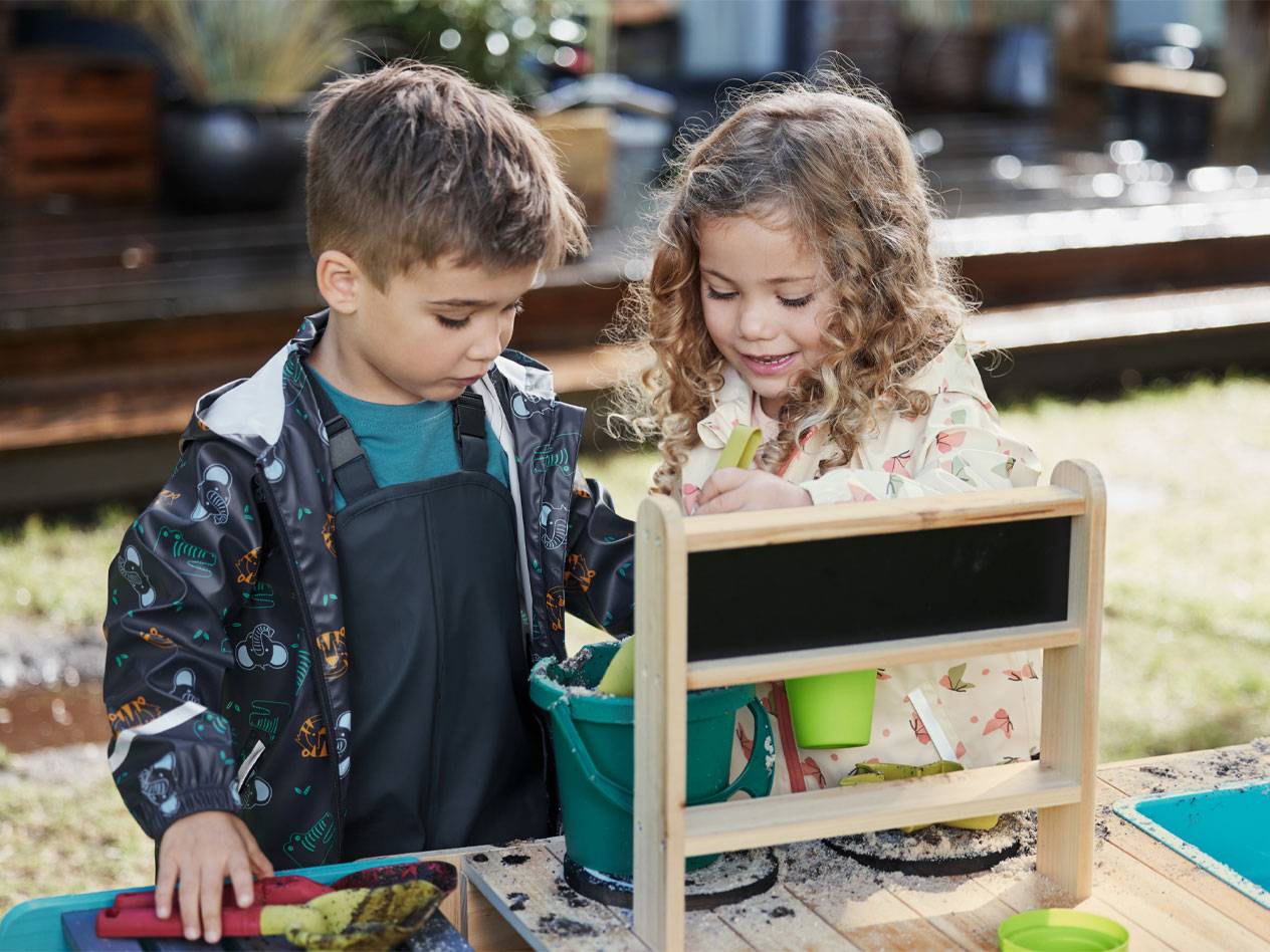 Two children in rain jackets and overalls play with sand at a play table.