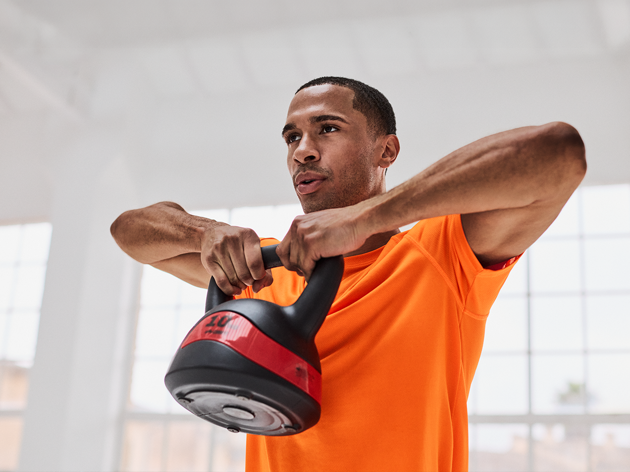 A man in an orange athletic shirt holding a black and red kettlebell.