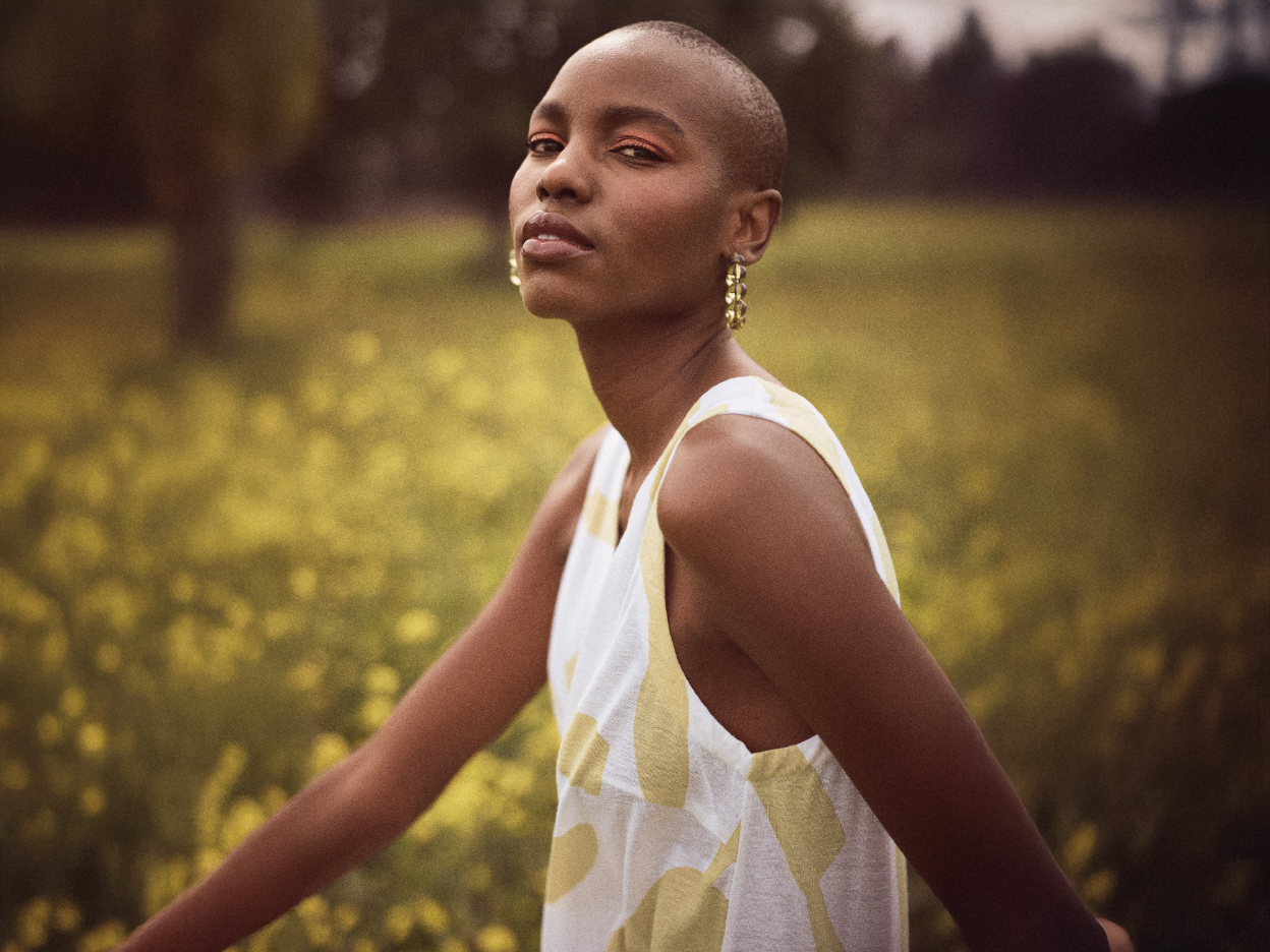 A woman with a shaved head wears a sleeveless, abstract-print dress in a field of yellow flowers.