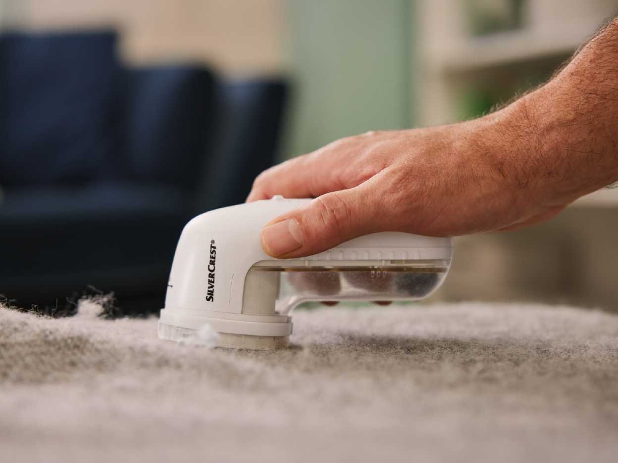 A hand uses a SilverCrest lint remover on a gray fabric, collecting lint.