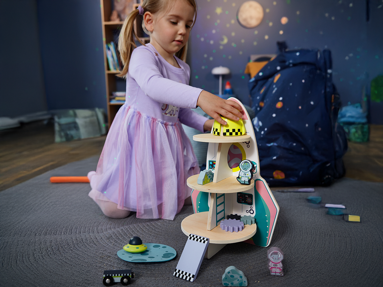 A young girl playing with a wooden rocket ship toy set on a rug.