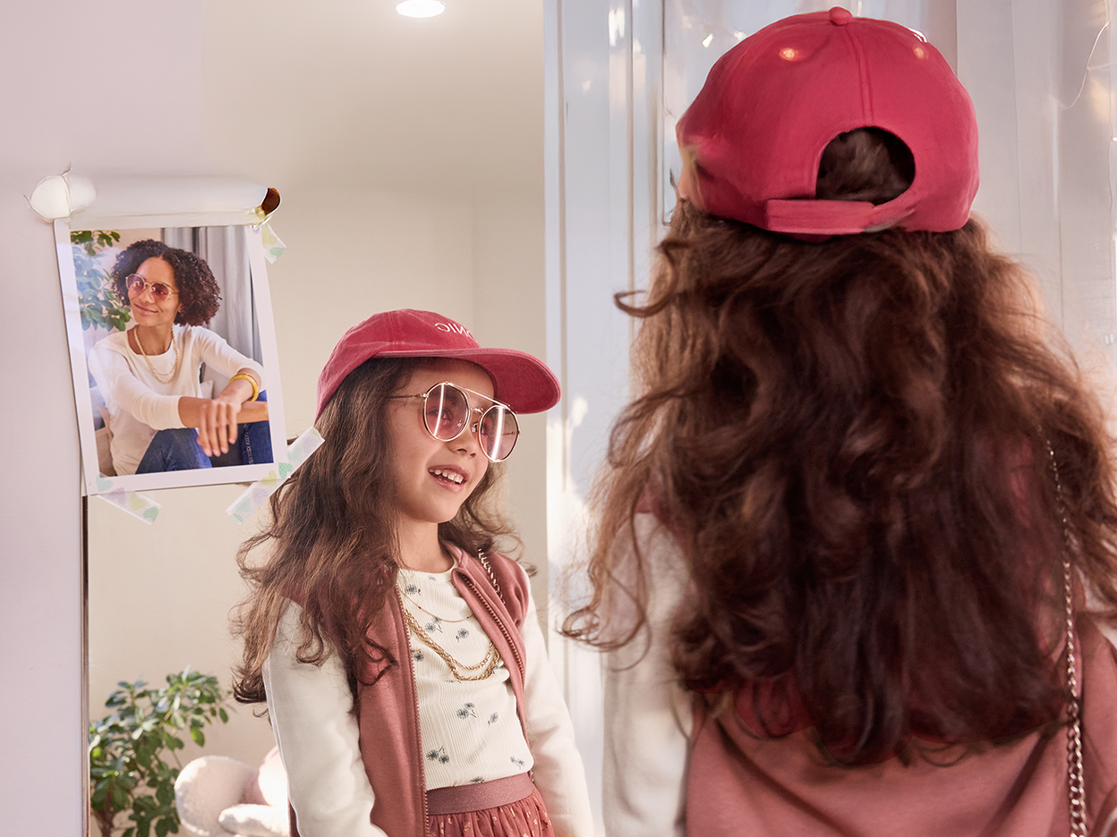 Girl in pink hat and sunglasses looking at her reflection in the mirror.