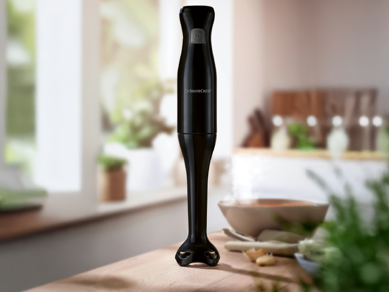 A black SilverCrest immersion blender stands on a wooden counter in a kitchen.