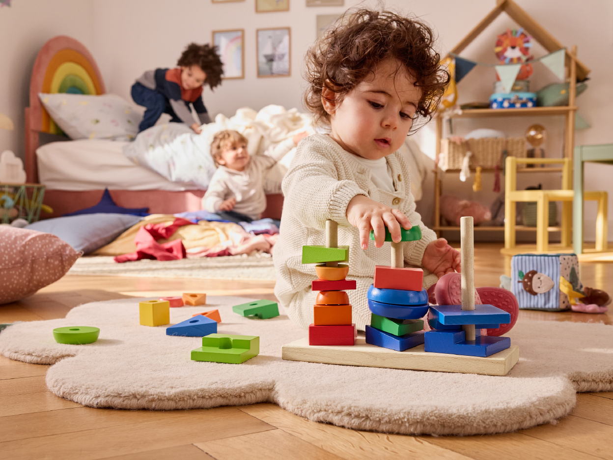 Toddlers playing with wooden stacking toys and blocks on a rug in a colorful bedroom.