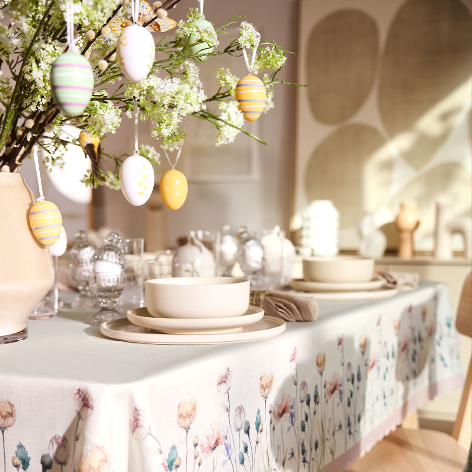 Easter table set with a floral tablecloth, ceramic dinnerware, and branches decorated with eggs.