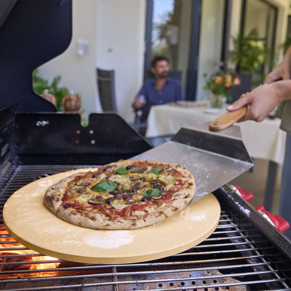 Pizza on a pizza stone on a grill, with a pizza peel and a man in the background.