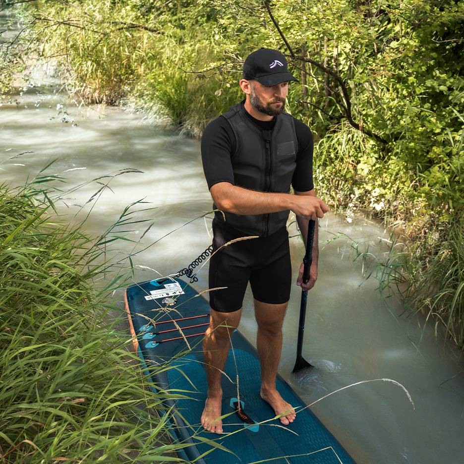 Man on a SUP board in a river, wearing a wetsuit and life vest.