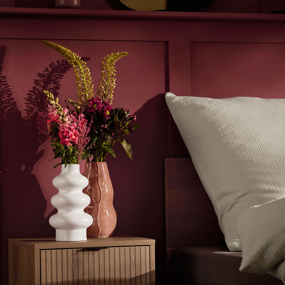 Two vases with flowers on a nightstand next to a bed with a white pillow.