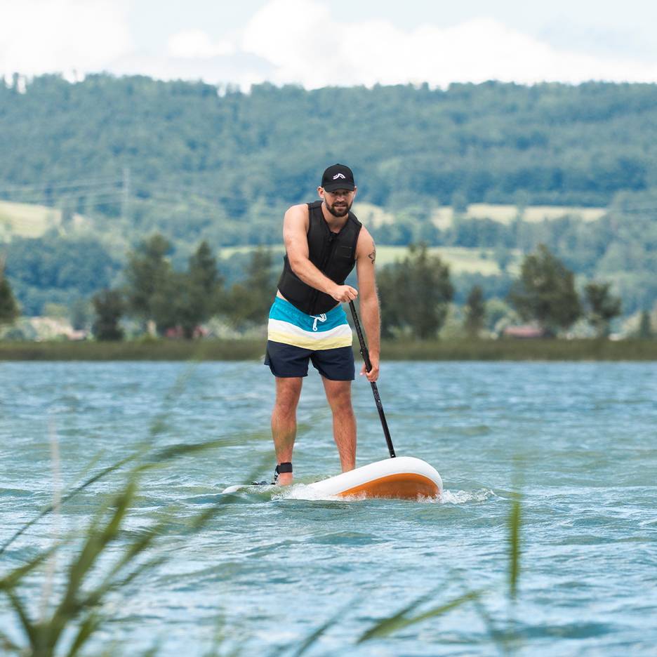 Man stand-up paddleboarding on a lake, wearing a life vest and swim shorts.