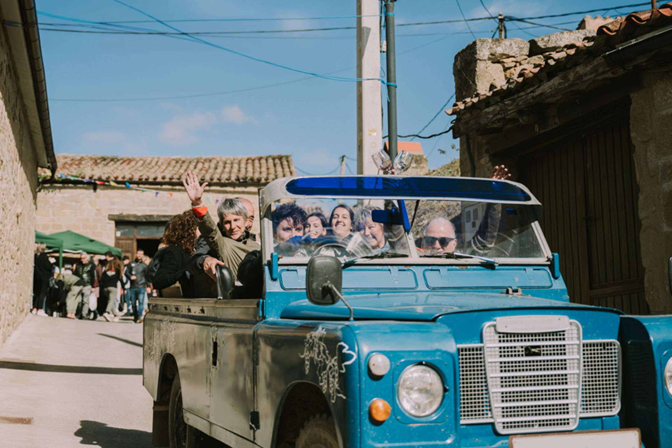 People in a blue vintage off-road vehicle driving through a village street.
