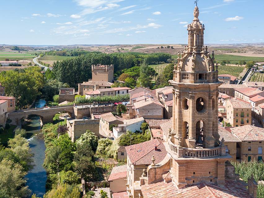 Aerial view of a historic town with a prominent church tower, river, bridge, and vineyards.