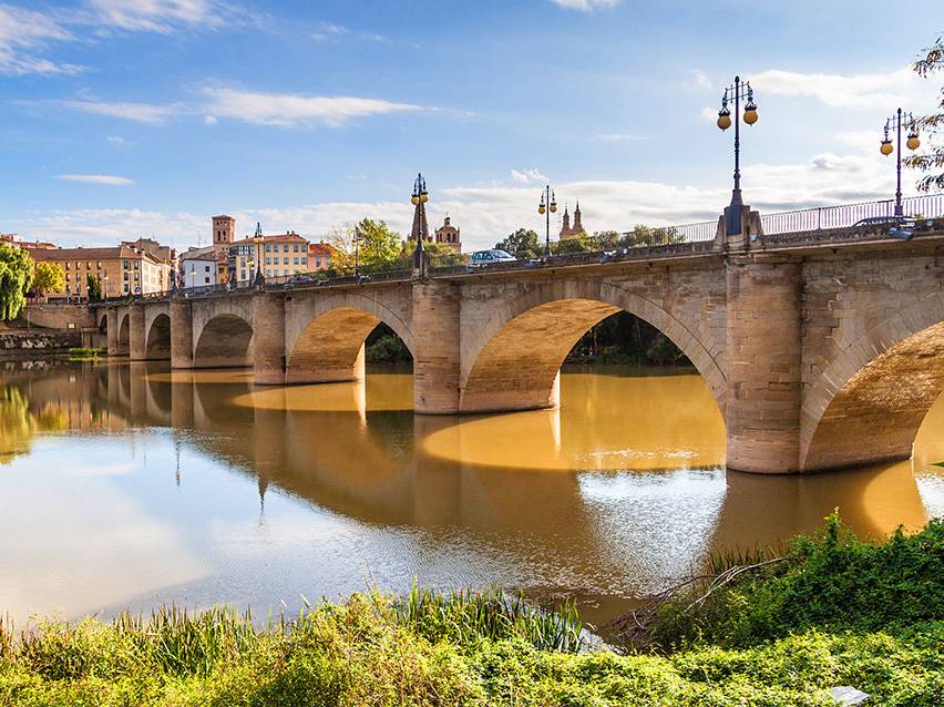 Stone arch bridge over a river with a town and blue sky in the background.