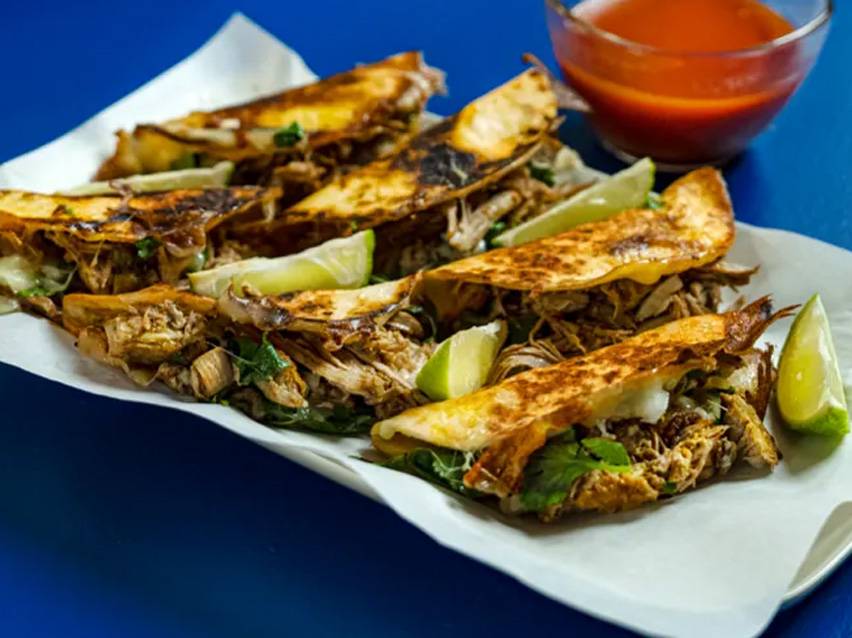 Crispy tacos with shredded meat, cilantro, lime wedges, and a side of red sauce.
