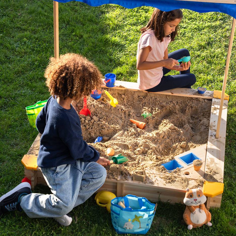 Two children playing in a sandbox with a canopy, surrounded by sand toys.