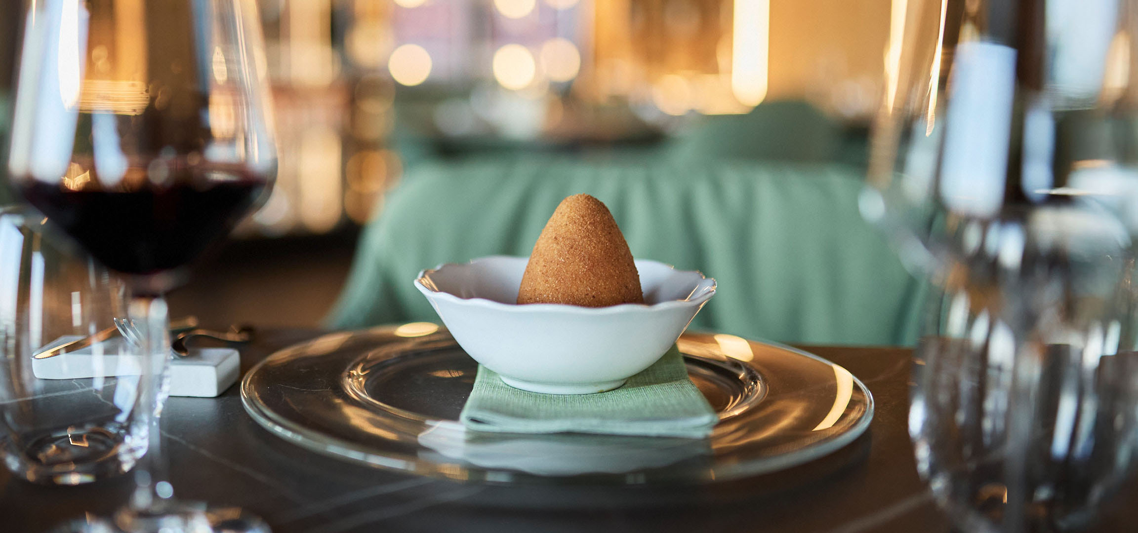 A golden conical dish in a white bowl, with red wine and cutlery on a table.