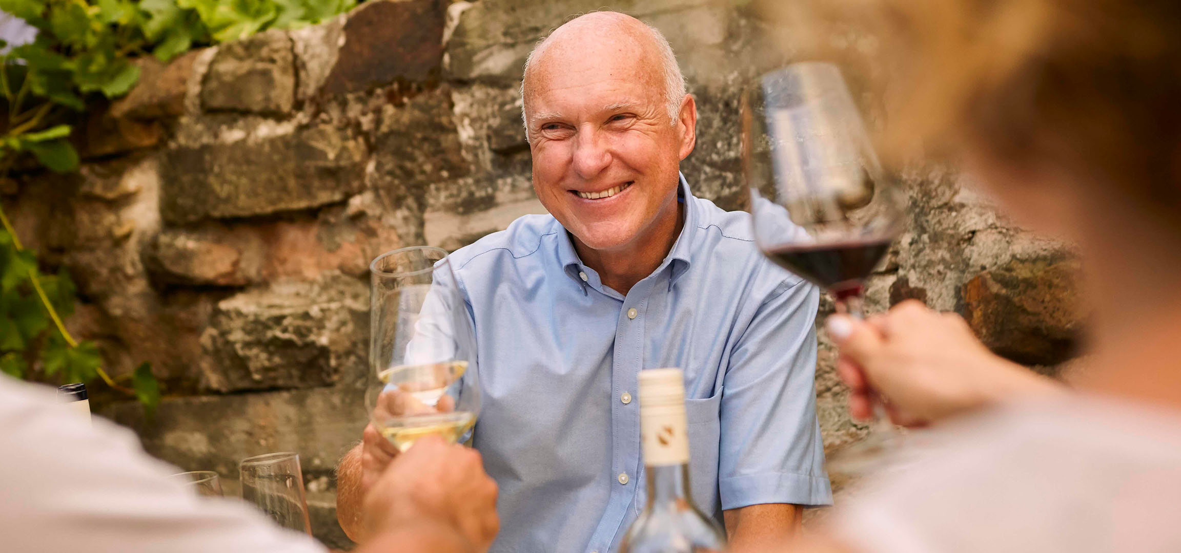 Smiling man enjoying red wine outdoors with a stone wall and greenery in the background.