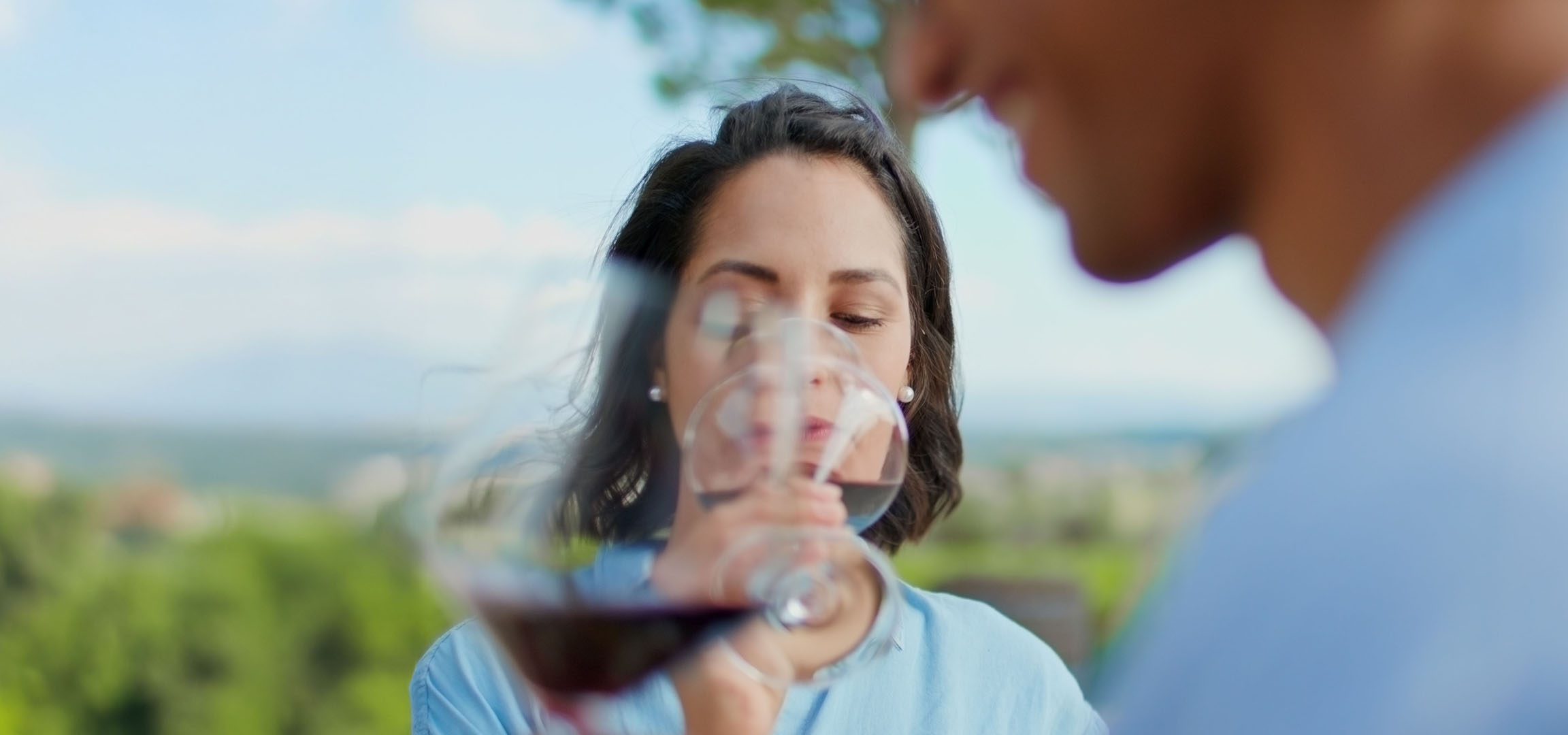 Woman enjoying red wine outdoors, man in background.