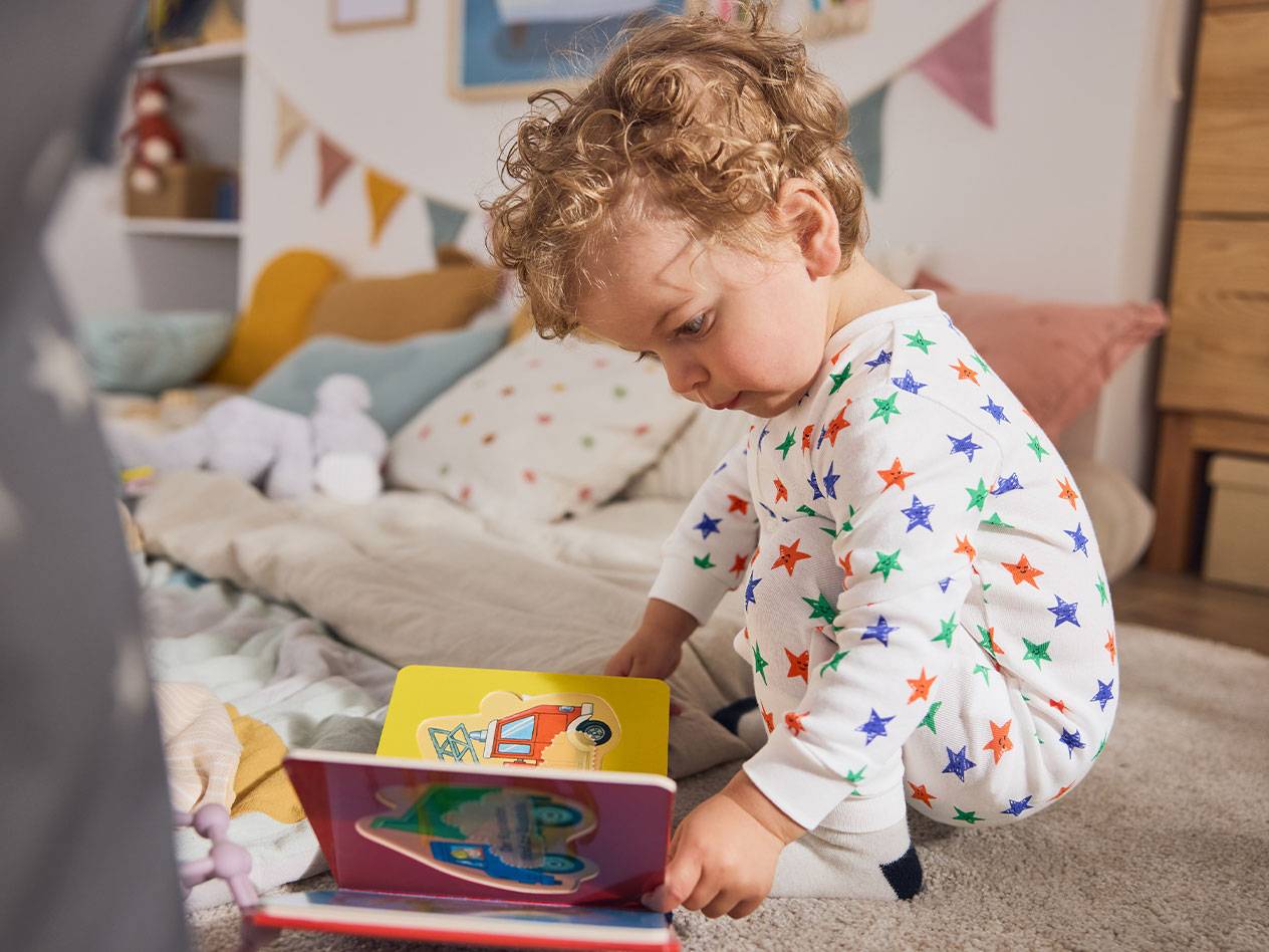 A toddler in star pajamas, sitting on the rug, reading a book.