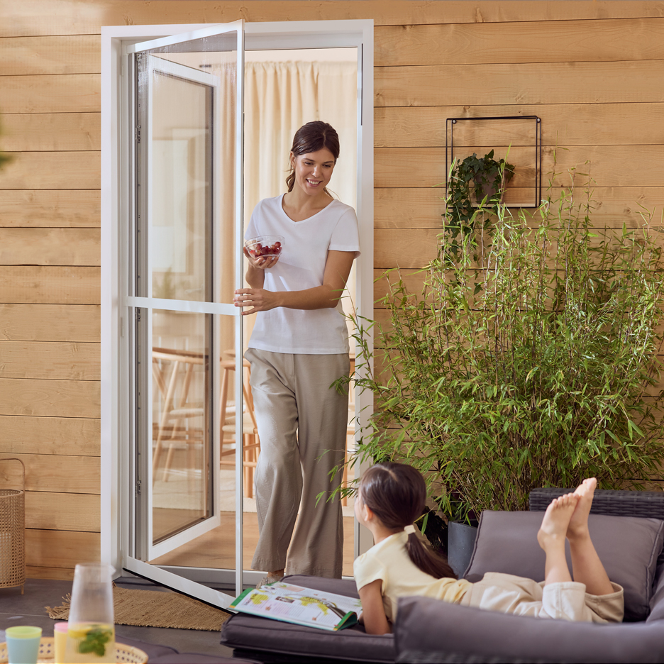 Woman with berries entering through a fly screen, child reading on a patio sofa.