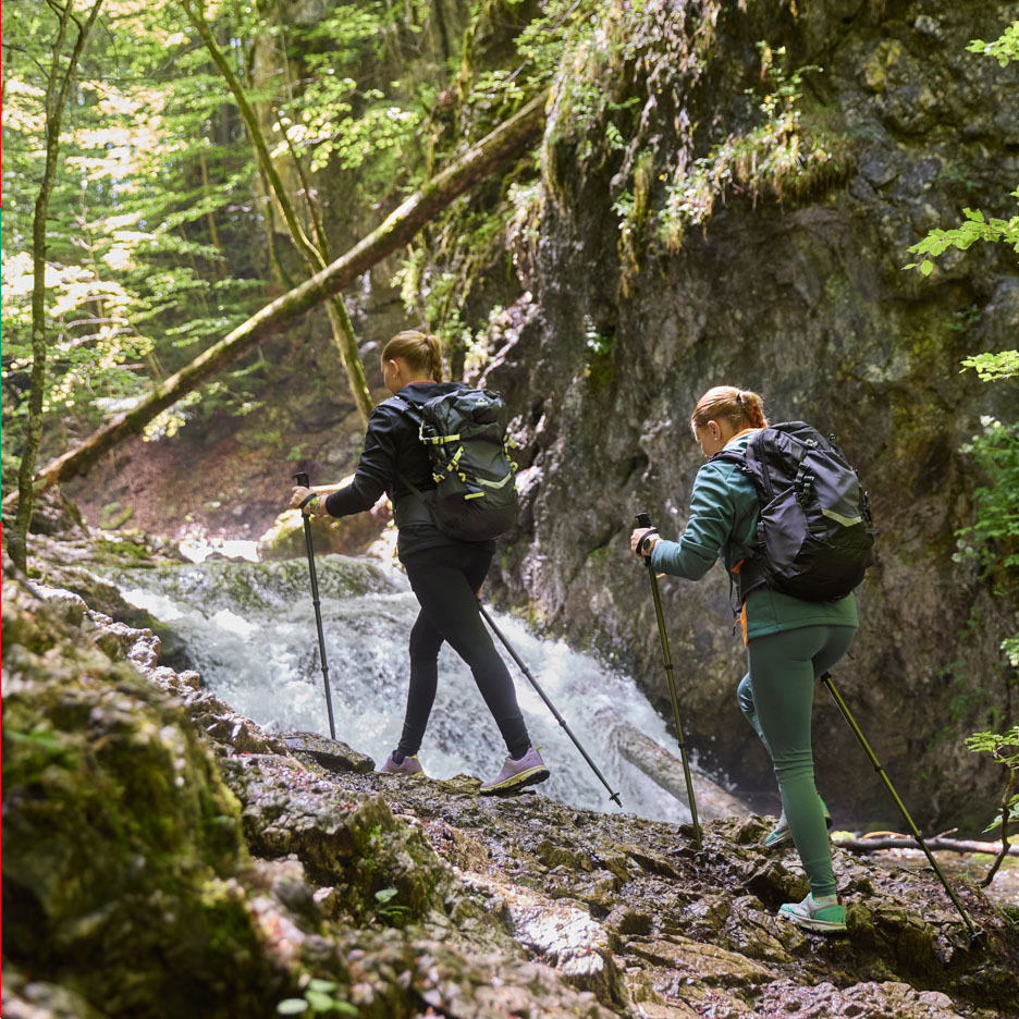 Two women with backpacks and trekking poles hike by a waterfall.